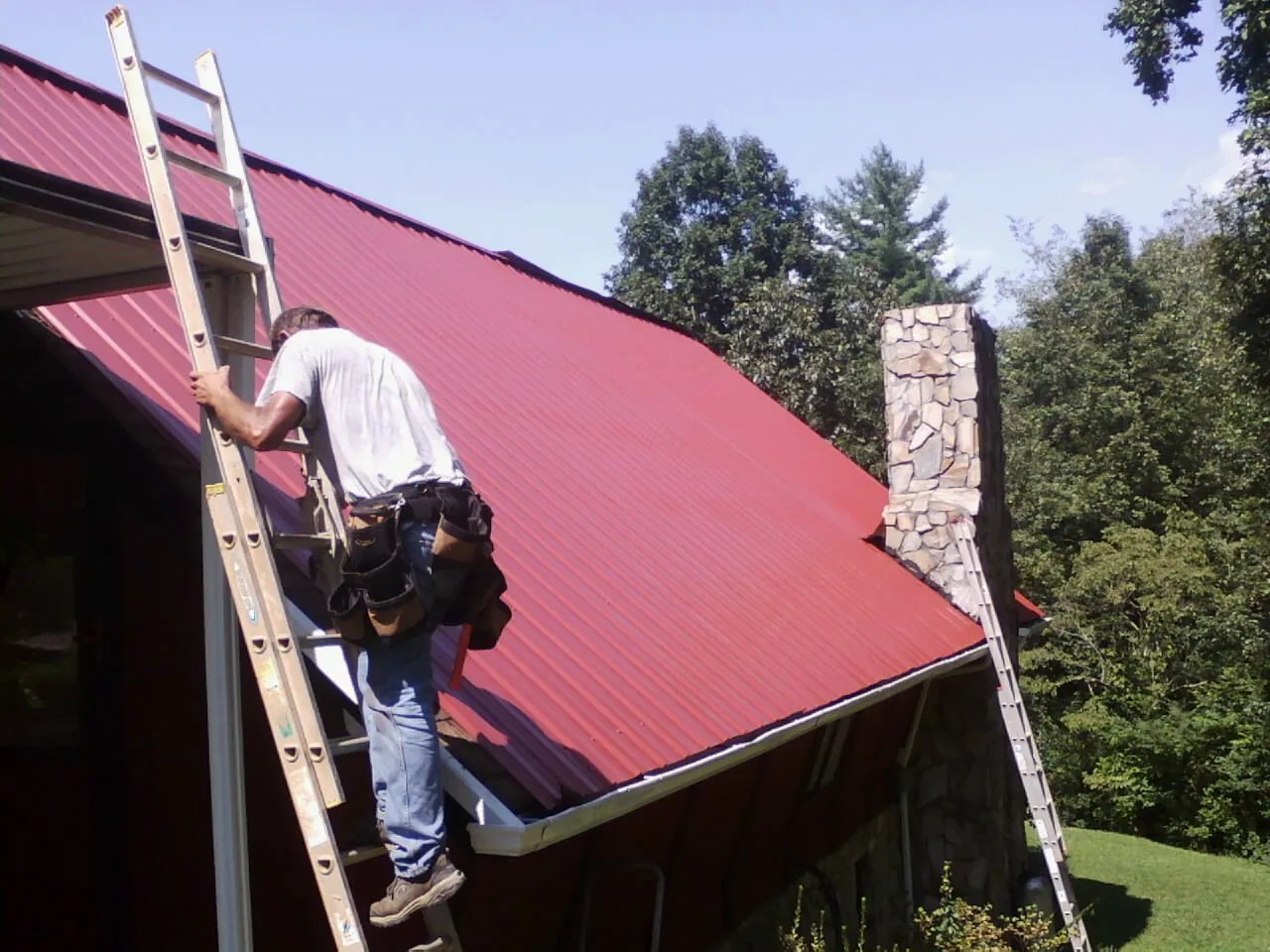 Roofer on a ladder beside a red metal roof and stone chimney — Soffit & Fascia Repair by Roofing Brothers in Signal Hill